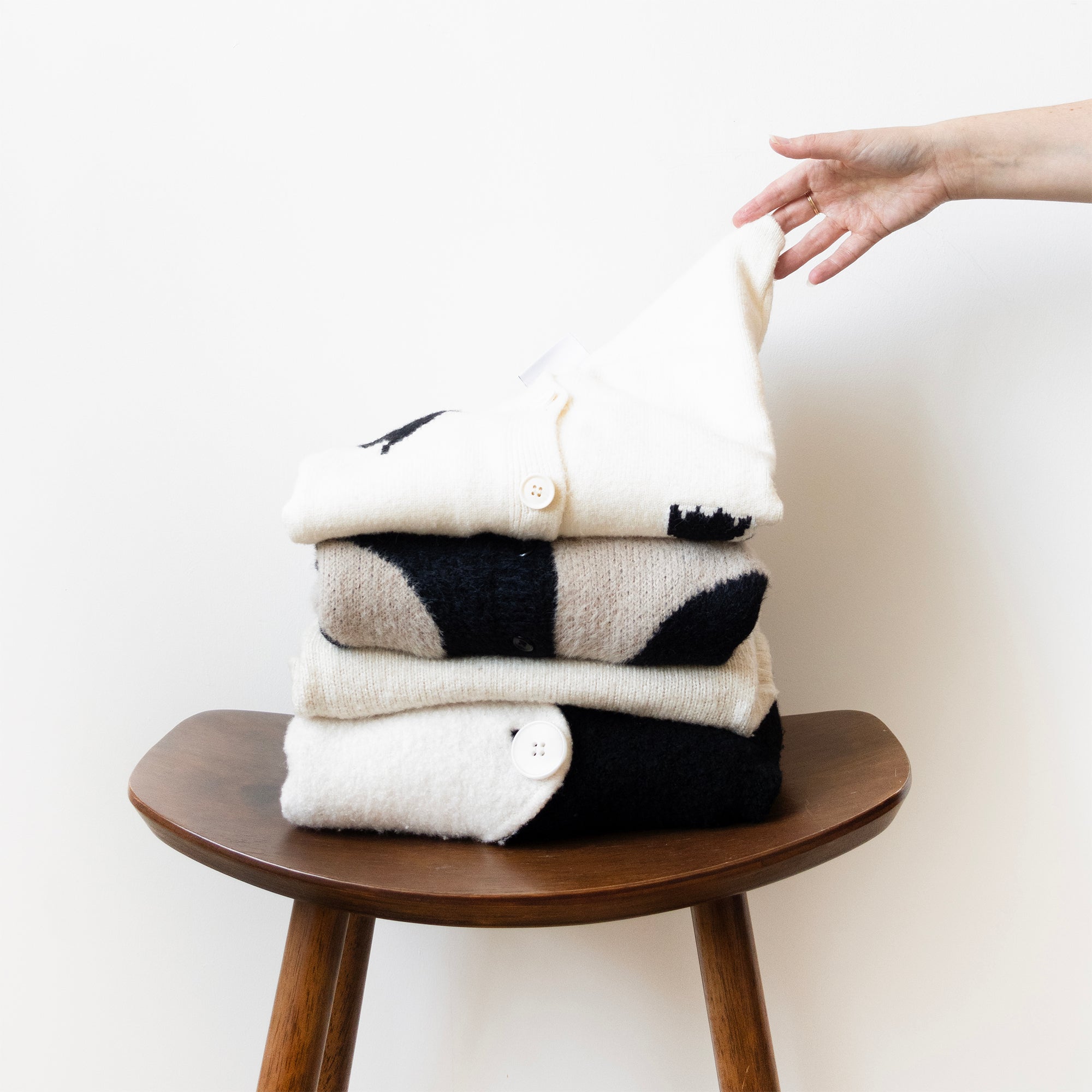 Stack of folded sweaters on a wooden stool with a hand reaching out.