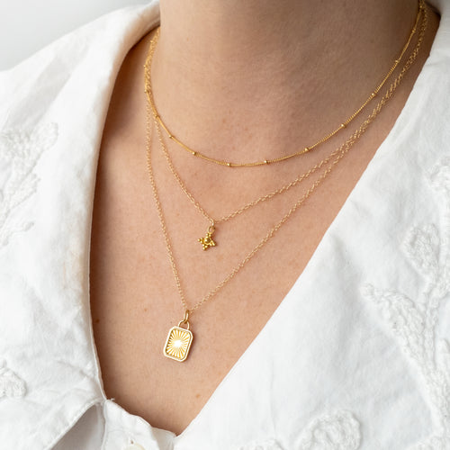 A close-up of a person's neck and chest wearing three layered gold necklaces over a white embroidered blouse. The necklaces include a beaded choker, a delicate chain with a small butterfly pendant, and a fine chain with a rectangular sunburst pendant.