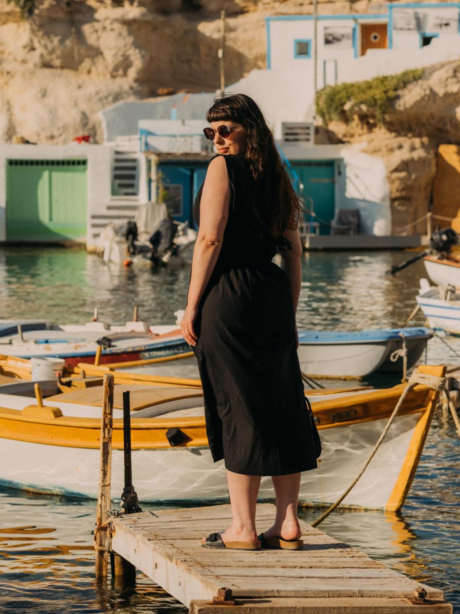 Woman in a black dress standing on a wooden dock by a body of water with boats and cliffs in the background.