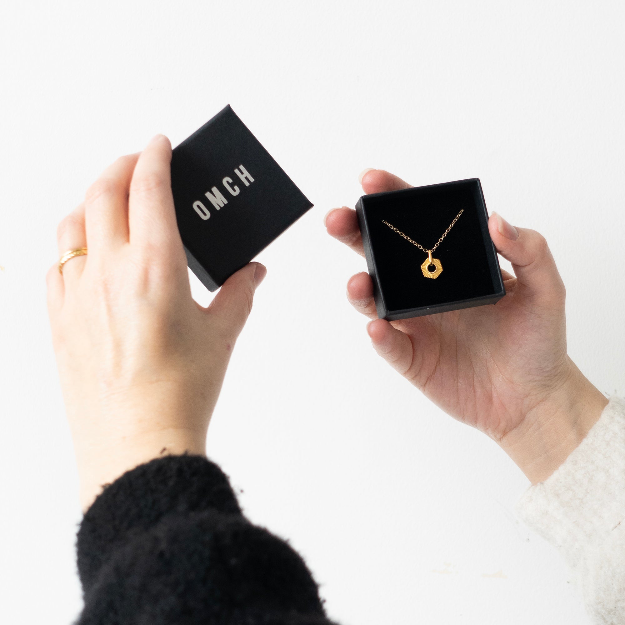 Person holding a gold necklace in a black box with 'OMCH' branding.