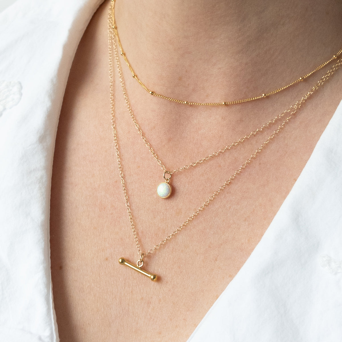 A close-up of a person's neck and upper chest wearing three layered gold necklaces: a delicate chain, a chain with a small white pearl pendant, and a chain with a small gold bar pendant, styled with a white top.